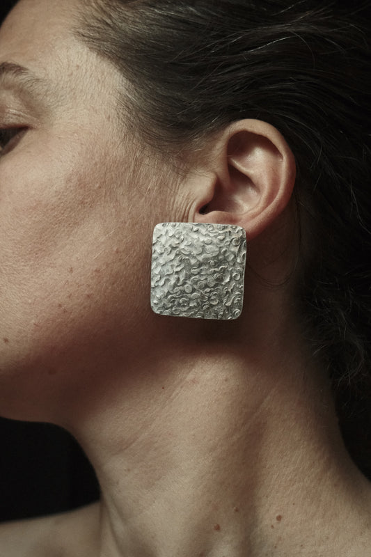 Boucles d’oreilles de forme carré, martelée donnant un aspect brut et irrégulier. Couleur argentée.