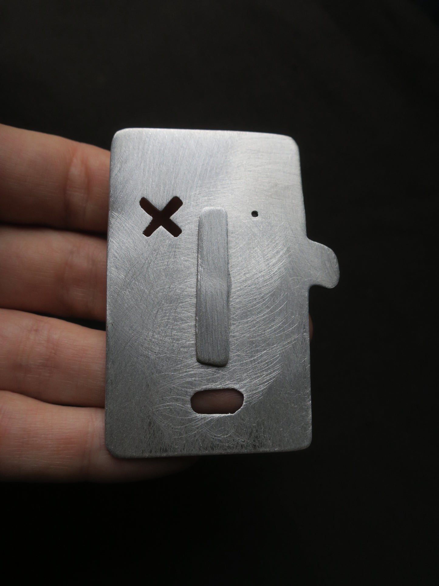 Broche en forme de visage caricaturale. D’inspiration industrielle, il s’agit d’une broche en aluminium brossé. Un oeil est un simple trou, l’autre une forme de croix évidée. Une asymétrie mutine est formée par l’oreille unique située sur le coté droit du bijou. 
